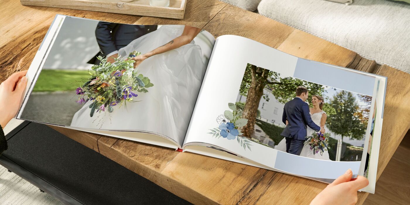 Offenes CEWE FOTOBUCH mit Hochzeitsfotos auf Holztisch, links Brautstrauß, rechts Brautpaar unter Bäumen