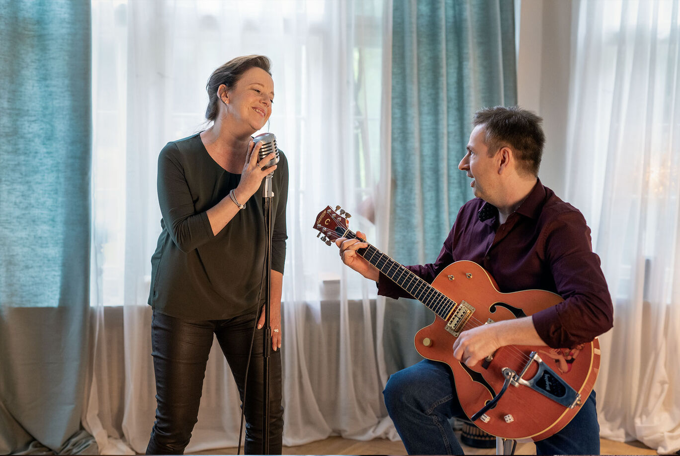 Ein Sänger trägt ein Mikrofon und singt. Ein Musiker mit einer Gitarre spielt dazu. Der Raum hat eine helle, freundliche Atmosphäre.