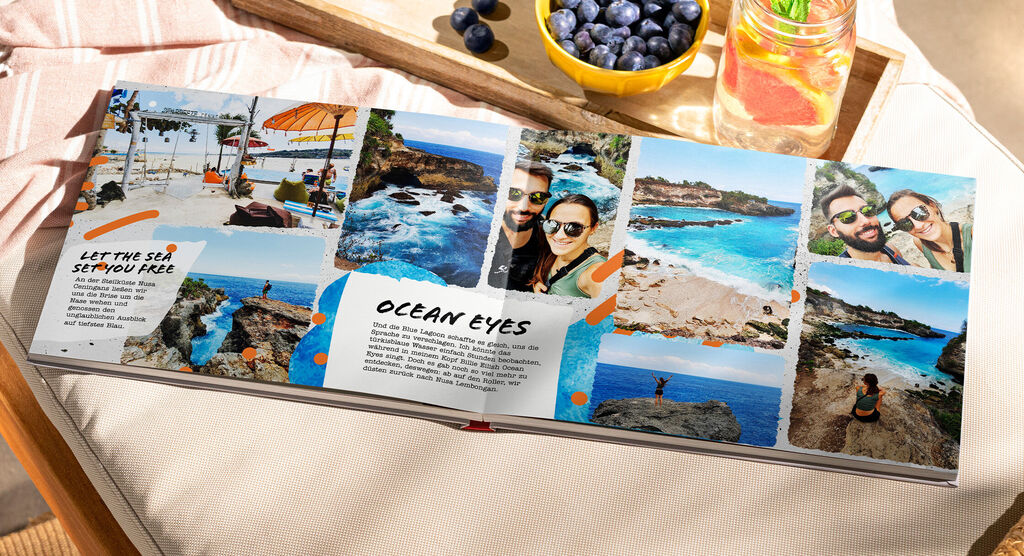 Ein geöffnetes Fotobuch mit schönen Urlaubsfotos vom Meer und Stränden, umgeben von frischen Früchten und Getränken
