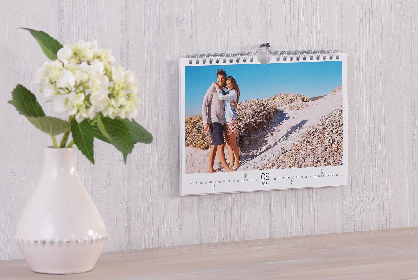 Ein CEWE Fotokalender hängt an der Wand. Er zeigt ein Paar, das sich umarmt, während sie am Strand stehen. Neben dem Kalender steht eine weiße Blumenvase mit einer Hydrangea.