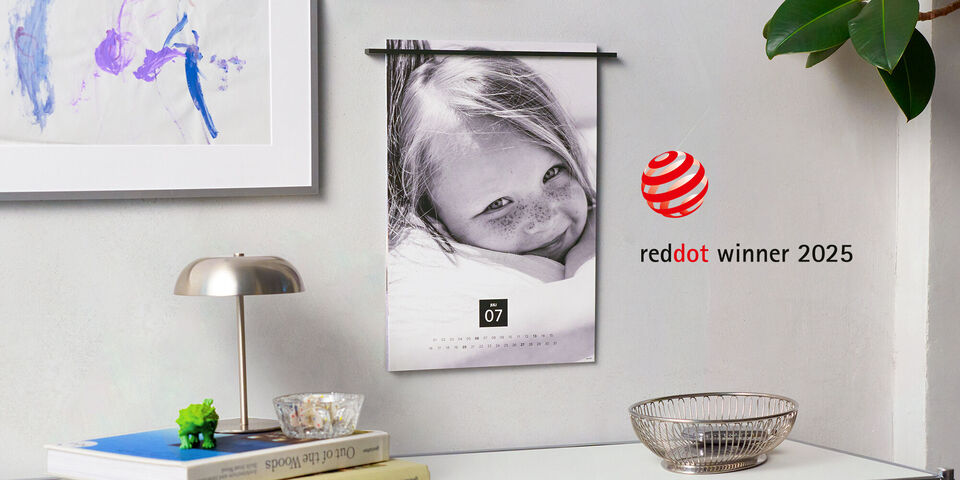 Ein schöner Kalender hängt an der Wand. Darauf ist ein lächelndes Kind mit Sommersprossen zu sehen. Das Bild lässt Freude und Lebensfreude ausstrahlen. Rechts neben dem Kalender ist eine moderne Tischlampe und eine Schale platzziert.