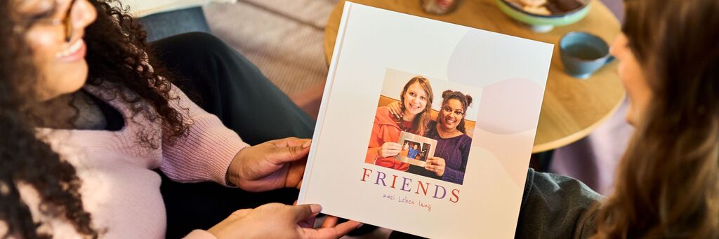 Zwei Personen halten ein fotografiertes Fotobuch mit dem Titel "Friends" in den Händen, auf einem Tisch stehen Tassen und eine Schüssel.