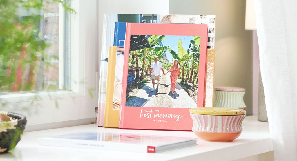 Auf einem Fensterbrett stehen mehrere Fotobücher. Auf dem ersten davon sieht man einen Mann und eine Frau im Sommerurlaub.