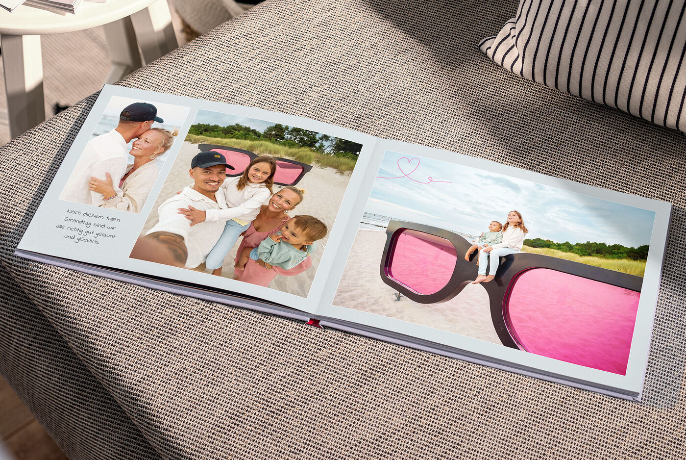 Auf einem Sofa liegt ein Fotobuch. Die Fotos zeigen eine Familie am Strand neben einer überdimensionalen Sonnenbrille mit pinken Gläsern.