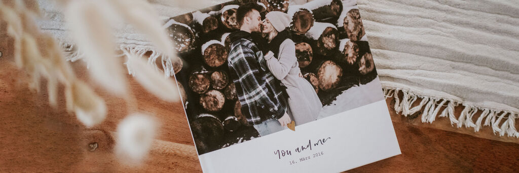 Ein Paar küßt sich glücklich vor einem Fotobuch mit romantischem Titel.
