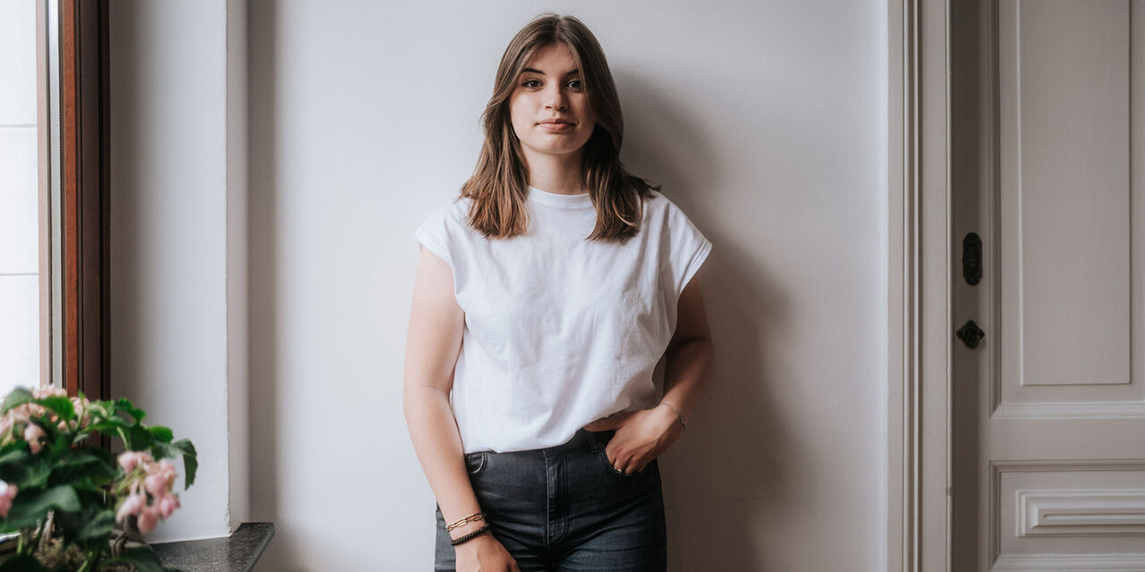 Eine Person steht lächelnd an einer Wand, trägt ein weißes T-Shirt und Jeans.