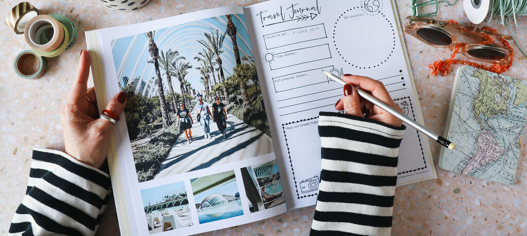 Hand einer Person hält einen Stift über ein offen liegendes Reisetagebuch, umgeben von einer Landkarte, Sonnenbrille und Klebebändern, eine wunderschöne Inspirationsquelle für Reiseerinnerungen