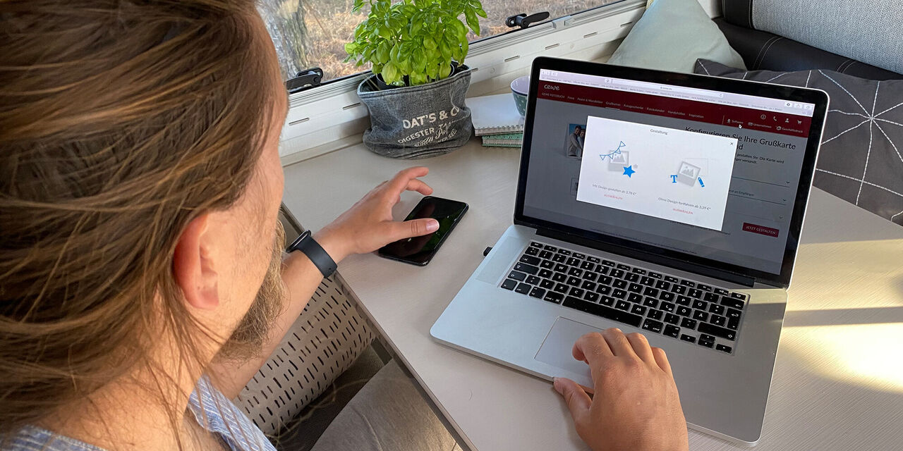 Mann mit Bart arbeitet am Laptop, hält ein Handy in der Hand.