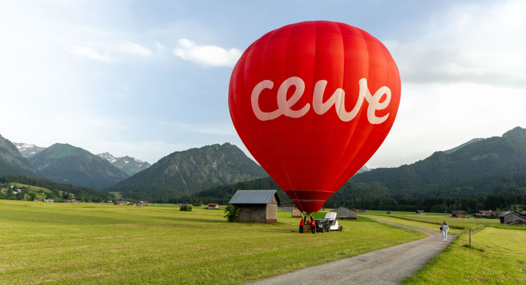 Ein roter Heißluftballon mit dem Schriftzug CEWE steht auf einer grünen Almwiese.
