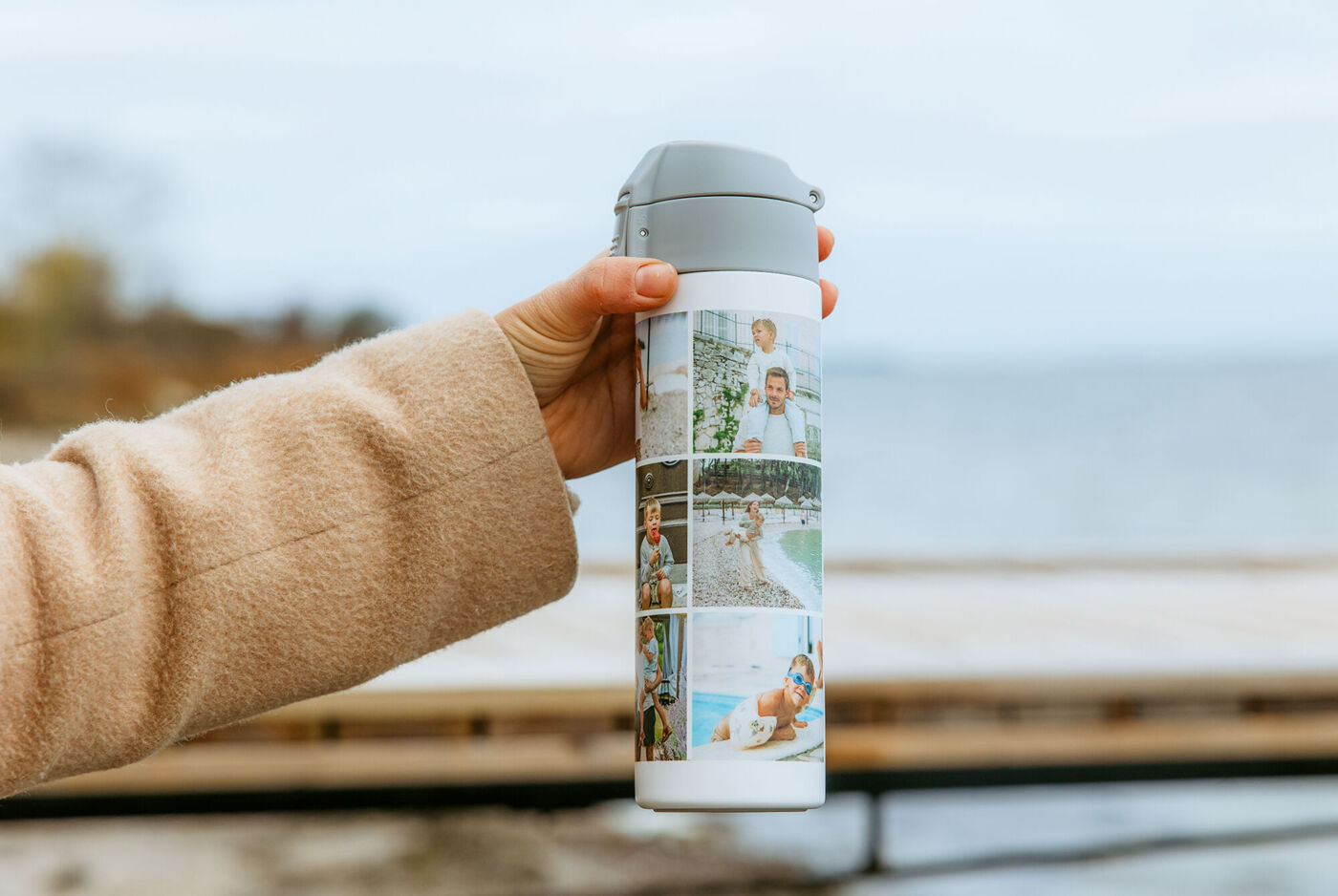 Hand hält eine liebevoll gestaltete Trinkflasche mit Fotocollage vor ruhiger See und weckt schöne Urlaubserinnerungen.