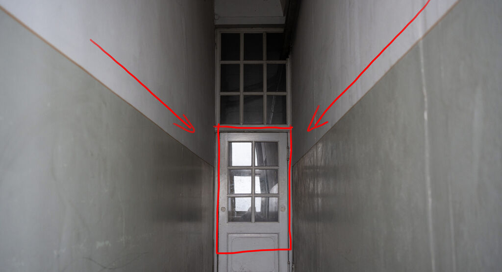 Eine weiße Tür mit Glasfenster in einem langen Flur. Auf beiden Seiten des Flurs sind graue Wände.