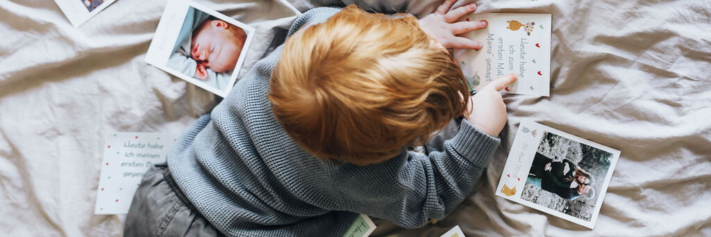 Ein kleiner Junge sitzt auf einem Bett und schaut sich Fotos an. Er hat rotbraune Haare und trägt einen grauen Pullover. Rund um ihn liegen viele bunte Fotografien.