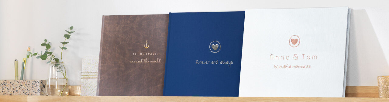 Drei wunderschöne CEWE Fotobücher stehen auf einem Regal. Verschiedene Designs in Braun, Blau und Weiß.