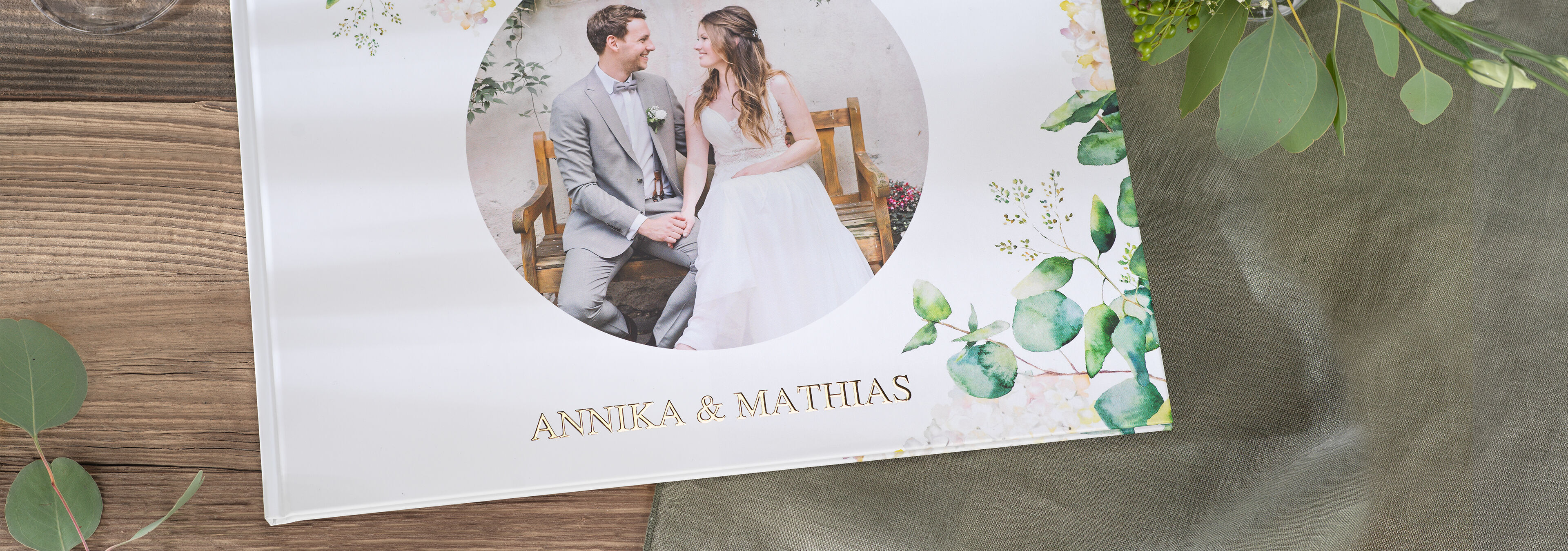 Ein schönes CEWE Fotobuch mit dem Titel Annika & Matthias auf einem Holztisch, umgeben von grünen Blättern.