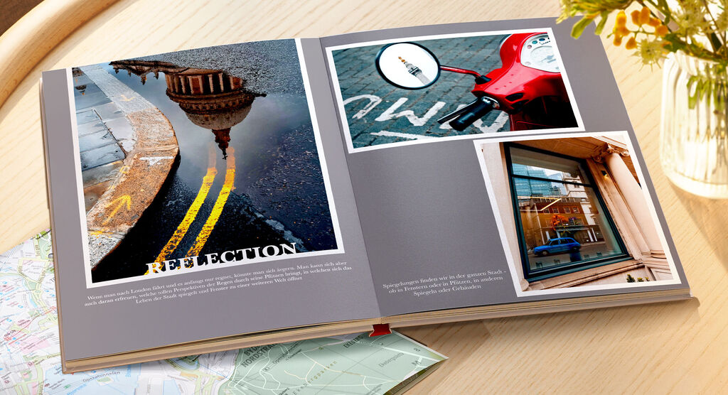 Das Bild zeigt ein CEWE FOTOBUCH, das auf einem Tisch liegt. Es enthält Fotos von reflektierenden Oberflächen, einer roten Vespa und einer Fensteransicht. Die Kulisse wirkt modern und urban.