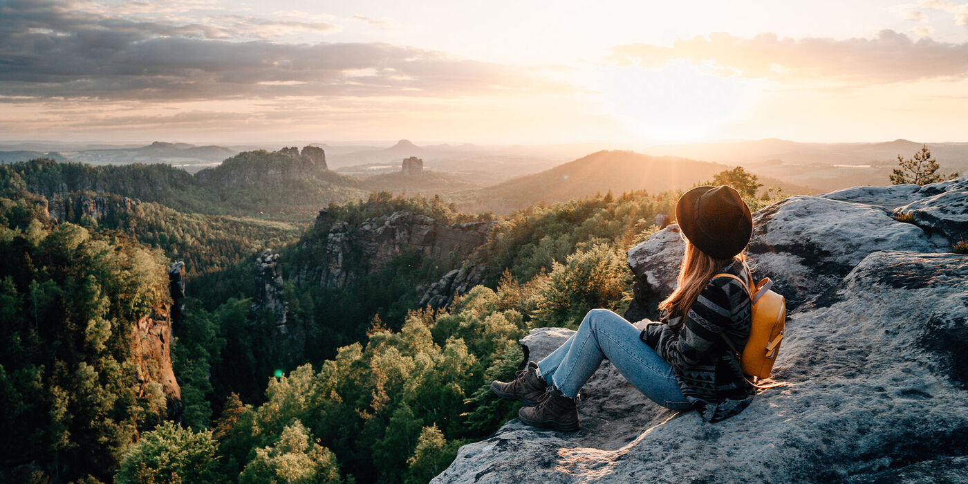 Frau sitzt auf einem Felsen und schaut auf eine wunderschöne Landschaft bei Sonnenuntergang. Bäume und sanfte Hügel sind im Hintergrund.