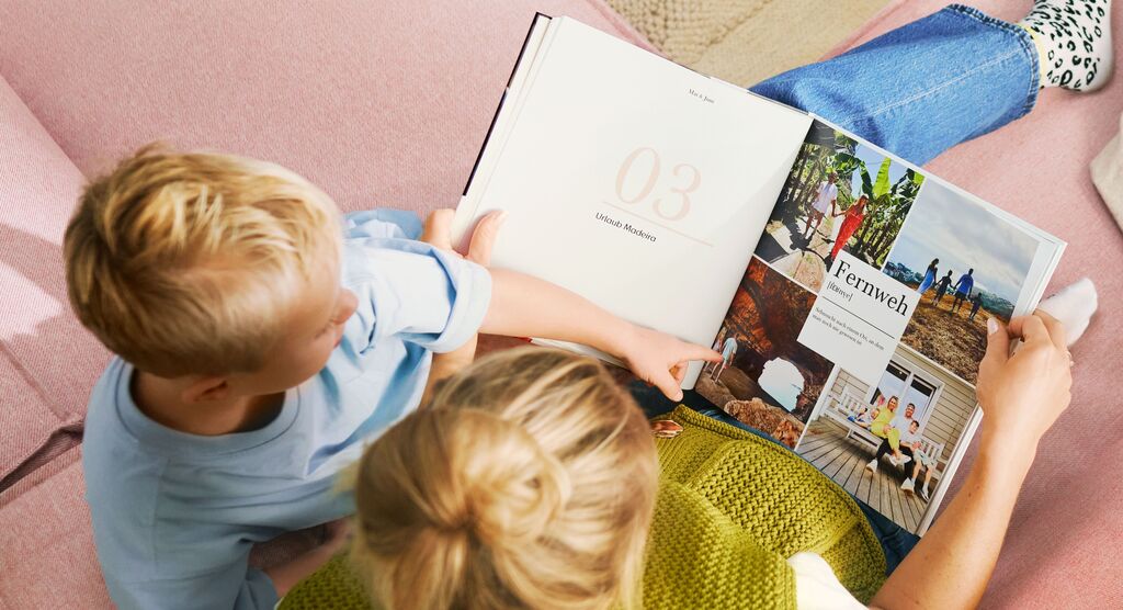 Eine Person liest mit einem Kind in einem CEWE FOTOBUCH auf einer pinken Couch. Auf einer Seite sind farbenfrohe Fotografie und Text zu sehen.