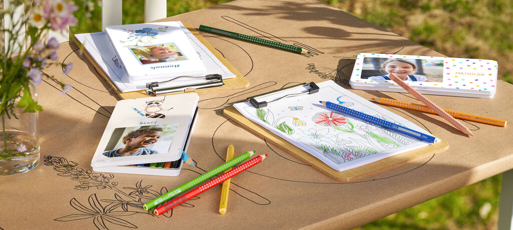 Bunte Bleistifte und Malhefte auf einem Tisch im Freien mit Fotografien und kreativen Materialien.