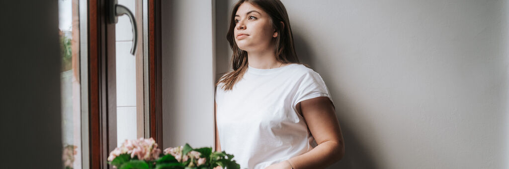 Eine Person steht vor einem Fenster und lächelt, während sie eine Blume in der Hand hält