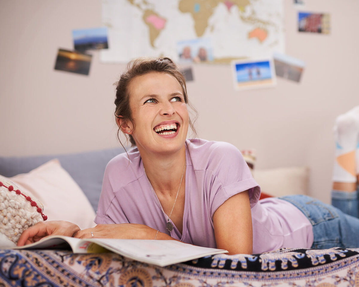 Frau liest in einem Magazin auf dem Sofa, umgeben von Postkarten und einer Weltkarte.