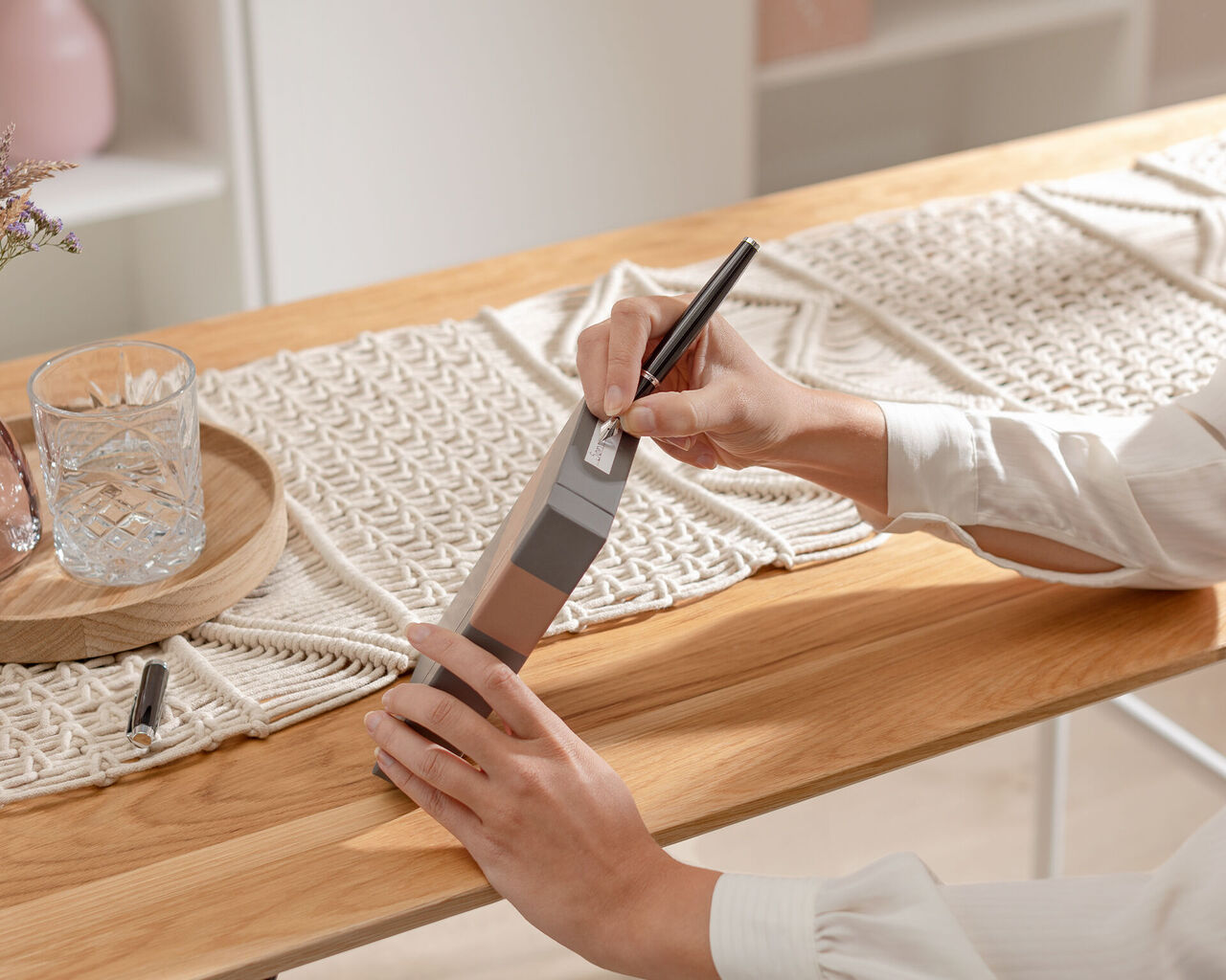Eine Person hält ein graues Notizbuch und schreibt mit einem schwarzen Stift darauf. Der Tisch ist aus Holz und hat dekorative Elemente.