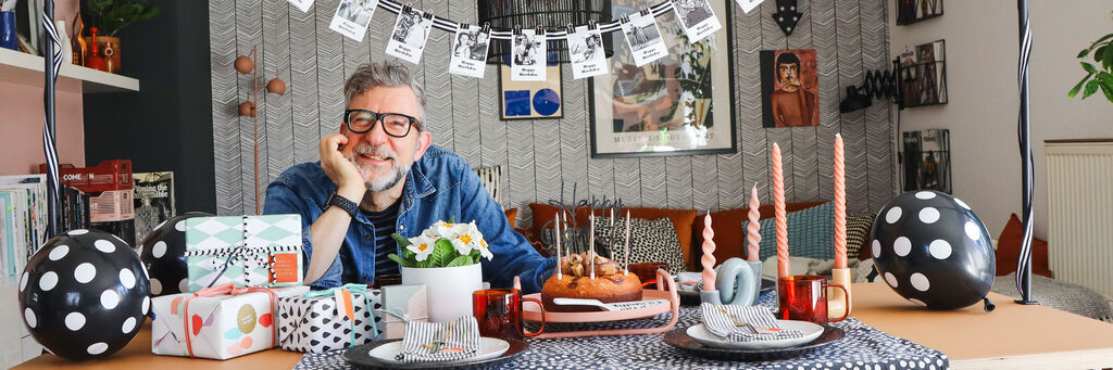 Ein Mann sitzt an einem festlich gedeckten Tisch mit Geschenken, Luftballons, Kerzen und Geburtstagsdekoration in einem Wohnzimmer