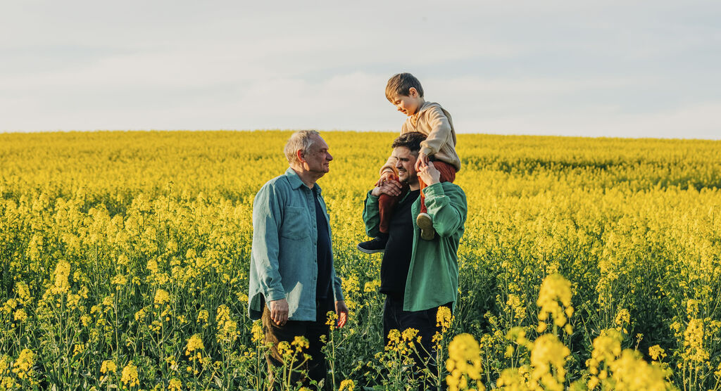 Eine fröhliche Familie steht auf einem gelben Blumenfeld. Die Eltern umarmen ihr Kind und genießen gemeinsam den Tag.