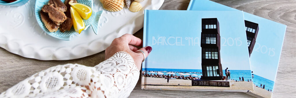 Eine Person hält ein CEWE FOTOBUCH vor einem Strand mit Menschen im Hintergrund.