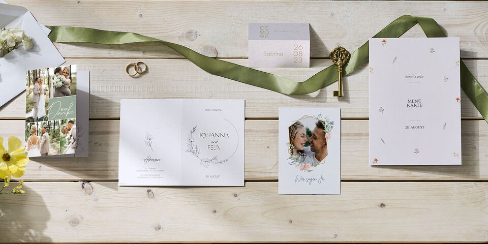 Hochzeitspapeterie mit Karten, Umschlag, grünem Band, Blumen, Ringen und Schlüssel auf hellem Holztisch arrangiert.