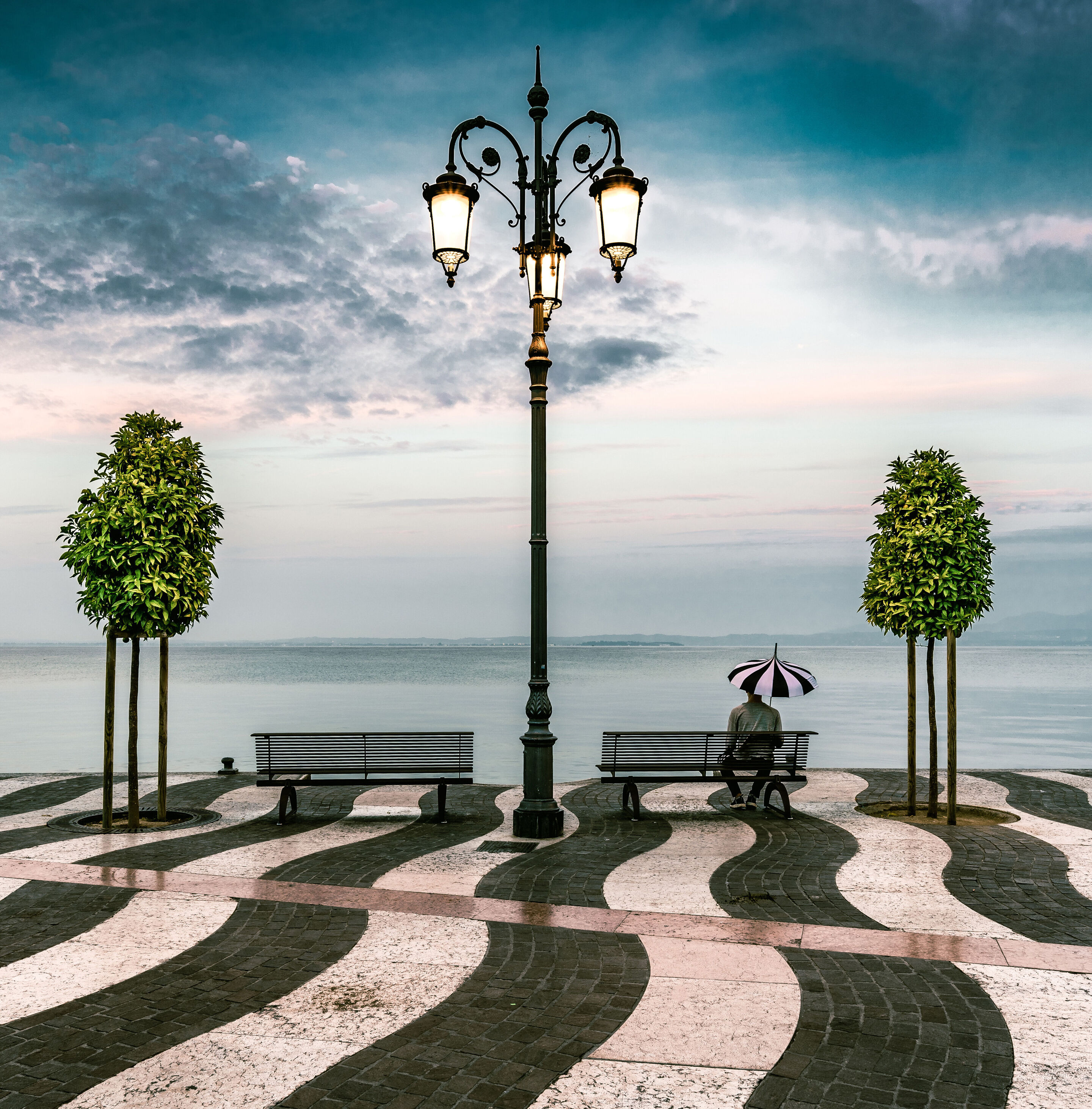 Auf einer schwarzweiß gepflasterten Seepromenade stehen zwei Bäume und zwei Bänke. Auf einer Bank sitzt ein Mann mit einem Sonnenschirm.