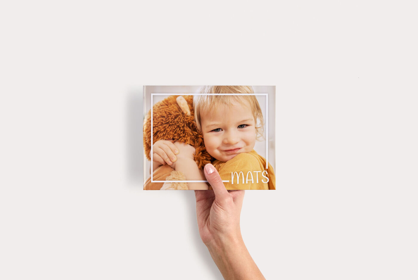 Hand hält ein CEWE FOTOBUCH Compact Panorama mit Kind und Teddybär auf dem Cover, minimalistischer Rahmen um das Foto
