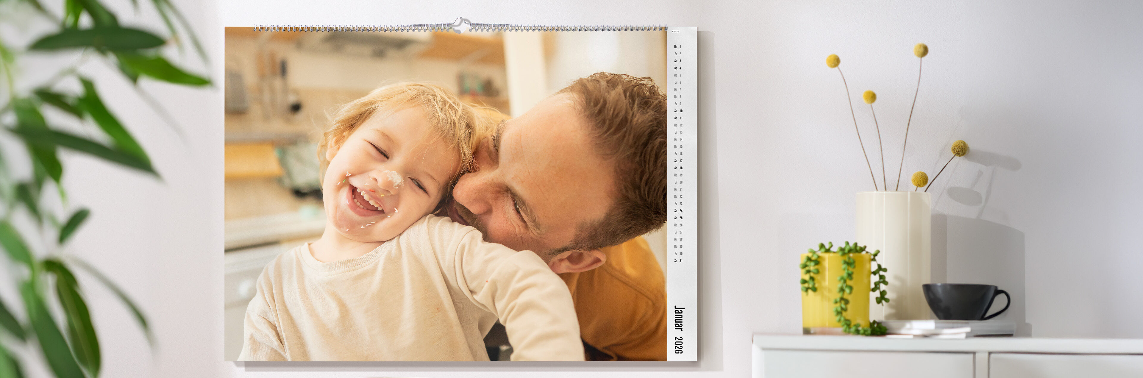 CEWE Fotokalender mit Familienfoto hängt an einer hellen Wand, daneben eine Kommode mit Vase, Pflanze und Tasse
