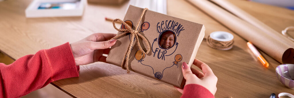 Eine Frau hält ein verpacktes Geschenk in den Händen. Eine Aussparung im Geschenkpapier zeigt einen Teil des Foto dahinter.