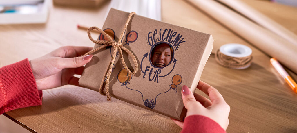 Eine Frau hält ein verpacktes Geschenk in den Händen. Eine Aussparung im Geschenkpapier zeigt einen Teil des Foto dahinter.