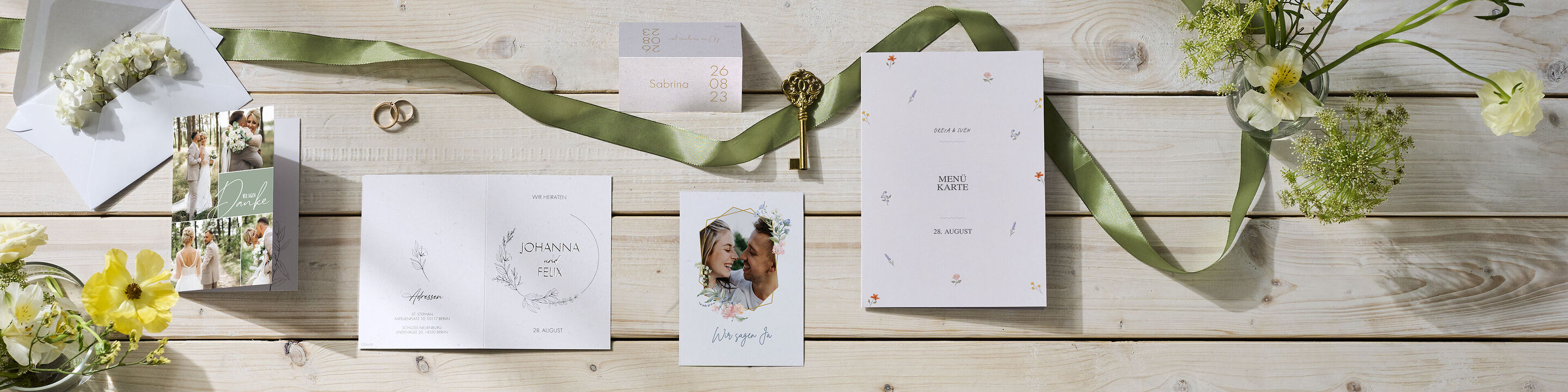 Hochzeitspapeterie mit Karten, Umschlag, grünem Band, Blumen, Ringen und Schlüssel auf hellem Holztisch arrangiert.