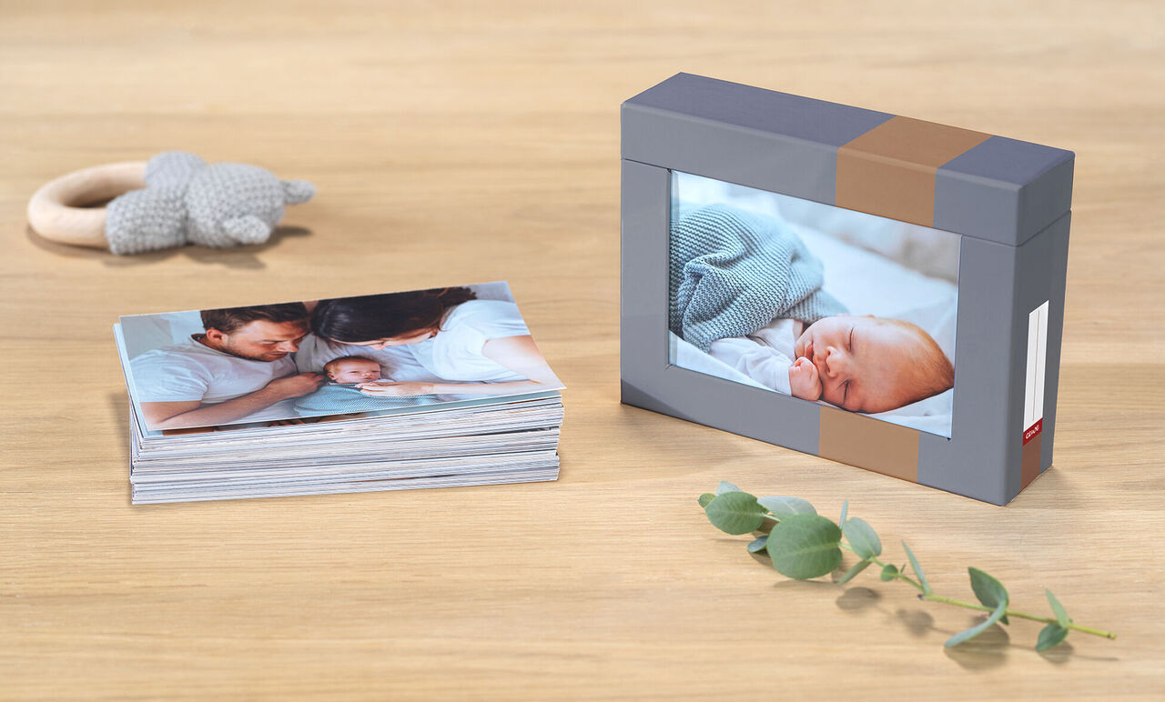 CEWE Fotobuch mit Fotos von einem Neugeborenen und Familie auf einem Holztisch, daneben eine Box mit einem Bild vom schlafenden Baby