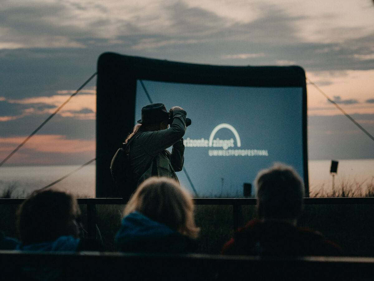 Ein Mann mit einer Kamera ist als Silhouette vor einer großen Leinwand zu erkennen.