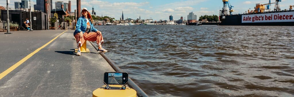 Person am Wasser sitzt auf einem Poller, sonniger Tag, Hamburg im Hintergrund, Handy mit Stativ vorne