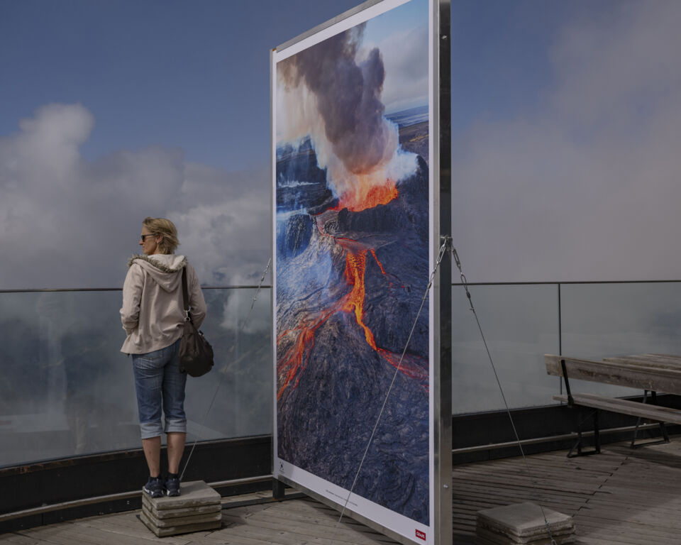 Ein großes Foto zeigt einen Vulkanausbruch. Es steht auf einer Terrasse. Daneben blickt eine Person in die Ferne.