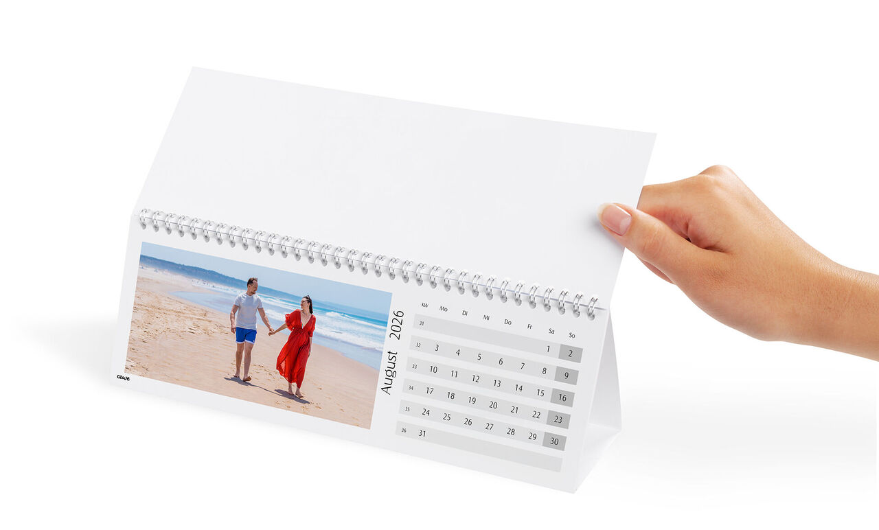 Eine Hand hält einen CEWE Fotokalender mit einem Paar am Strand. Der Monat August 2026 ist sichtbar