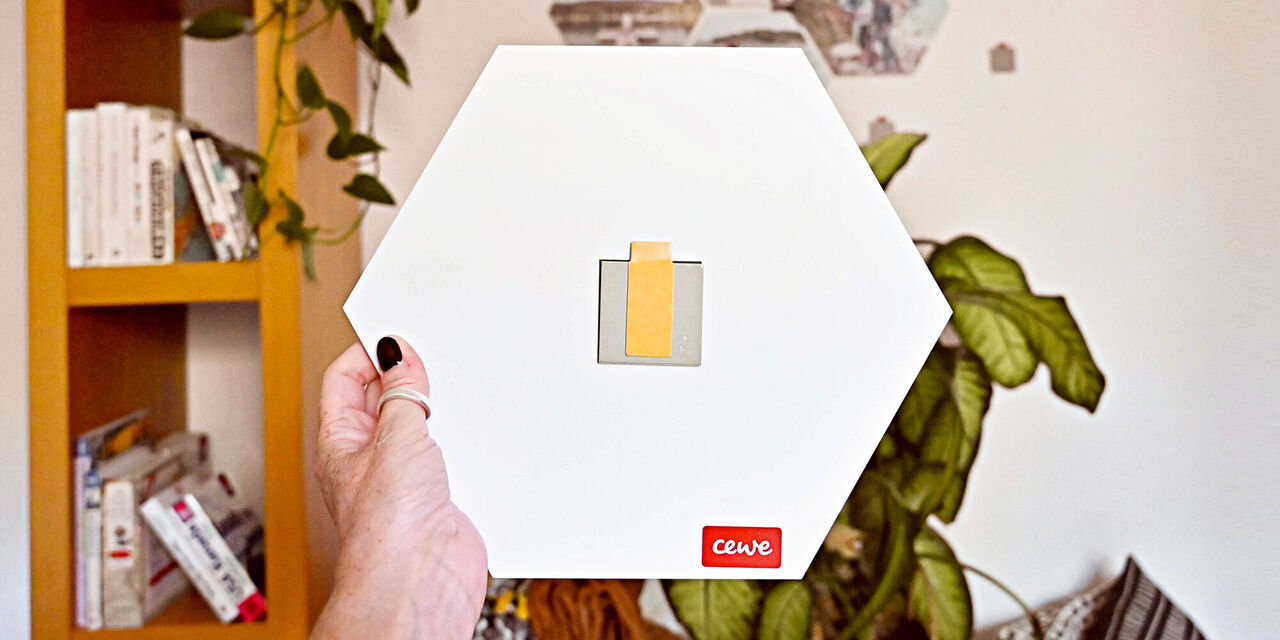 Eine Person hält ein hexagonales CEWE Fotobuch in der Hand, mit einem gelben Klebeband auf der Vorderseite.