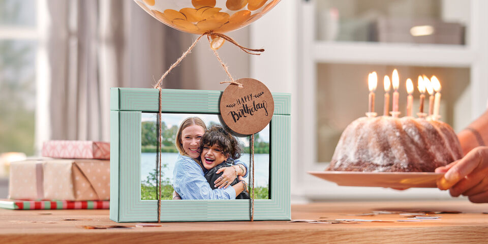 Gerahmtes Foto mit Geburtstagsmotiv vor Kuchen mit Kerzen und Geschenken im hellen Wohnzimmer.