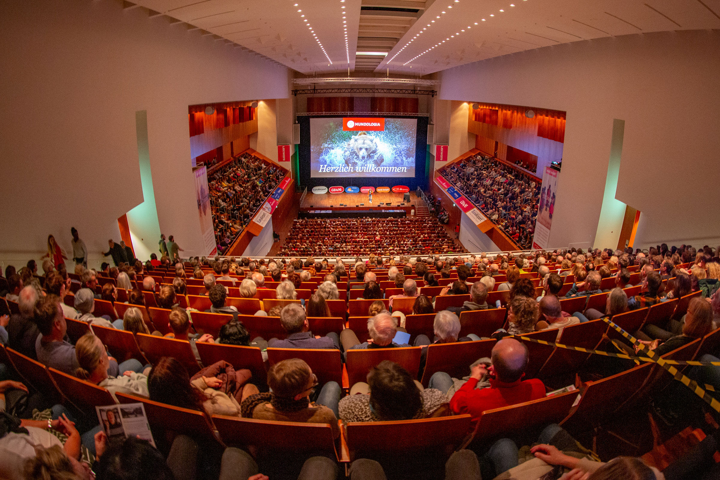 Ein vollbesetzter Konzertsaal, auf der Bühne ist eine große Leinwand mit einer Fotografie von einem Bären zu sehen.