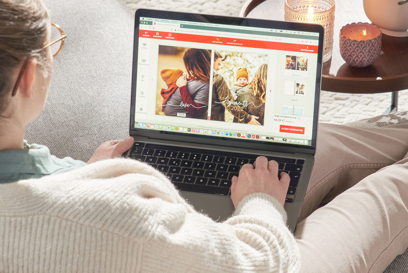 Eine Person sitzt auf einer Couch und schaut auf einen Laptop-Bildschirm. Dort sind zwei Fotobücher mit Babyfotos sichtbar. Der Raum ist gemütlich und warm mit einer Kerze.