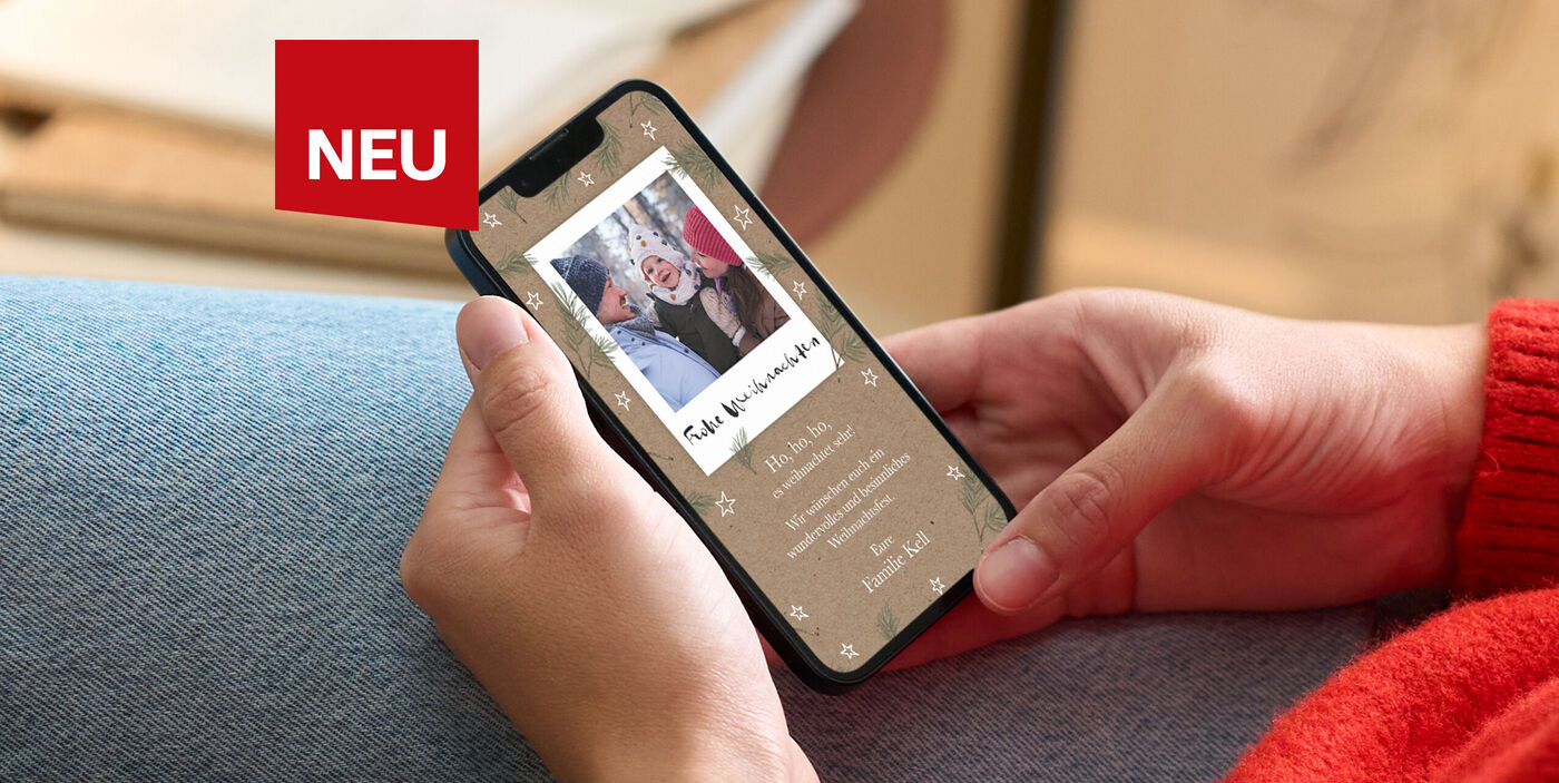 Frau in rotem Pullover sitzt gemütlich auf dem Sofa und liest eine digitale Weihnachtsgrußkarte auf ihrem Smartphone