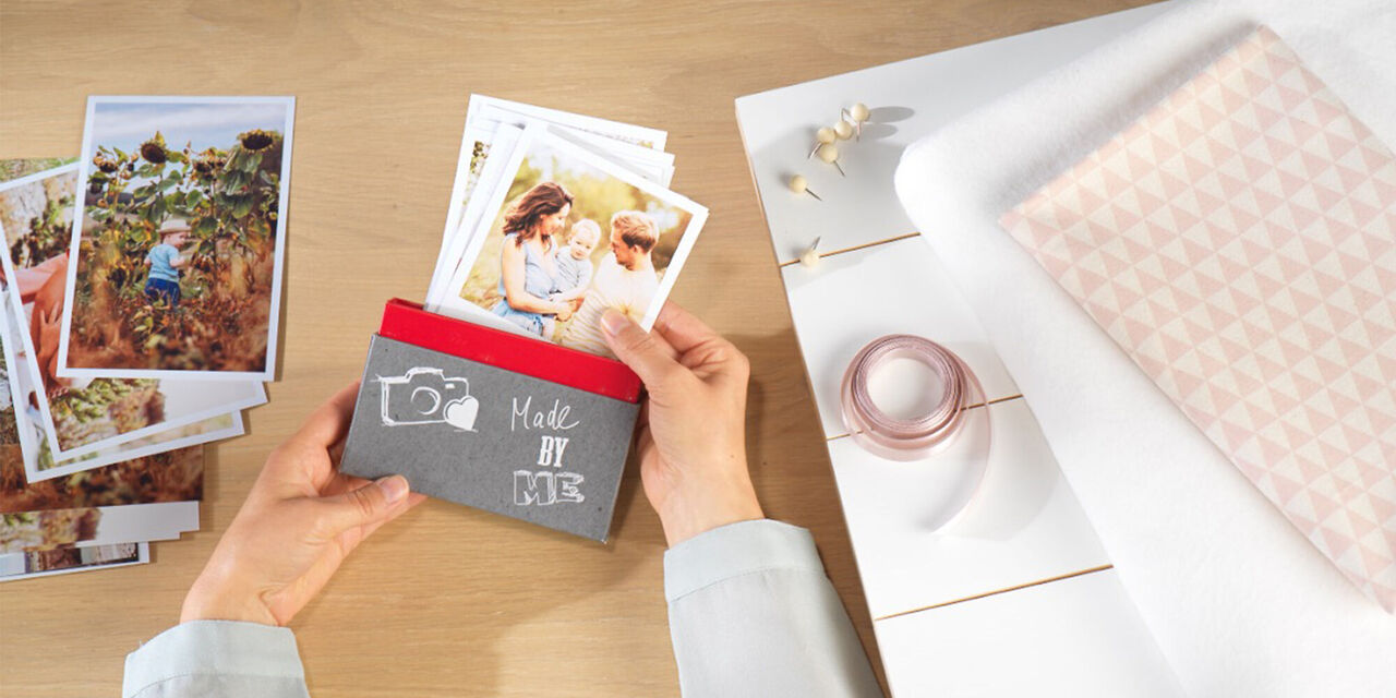 Eine Person hält eine kreative Fotoverpackung mit Bildern. Auf dem Tisch sind weitere Fotografien und Bastelmaterialien.