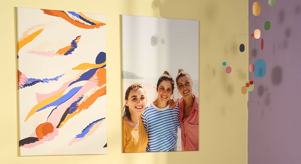 Zwei CEWE Wandbilder an einer gelben Wand, links mit buntem abstrakten Design, rechts mit einem fröhlichen Motiv