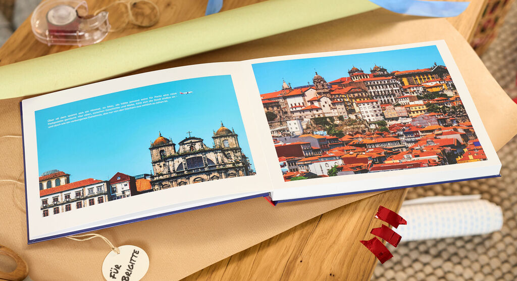Offenes CEWE FOTOBUCH mit Stadtansichten und roten Dächern vor blauem Himmel.