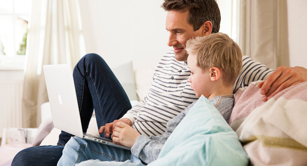 Ein Erwachsener und ein Kind sitzen gemeinsam auf einer Couch und schauen auf einen Laptop. Sie wirken entspannt und glücklich.